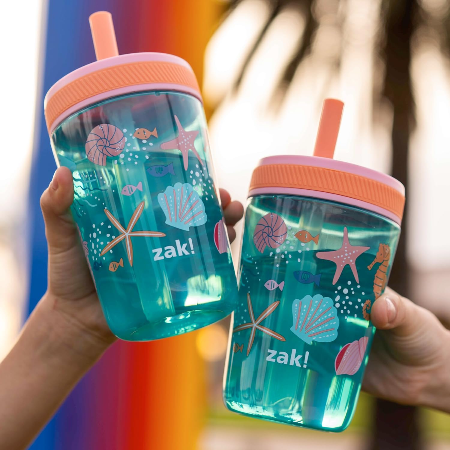 Two sea shells tumblers with pink lids and straw holders, featuring sea-themed designs, held by a person against a blurred outdoor background.