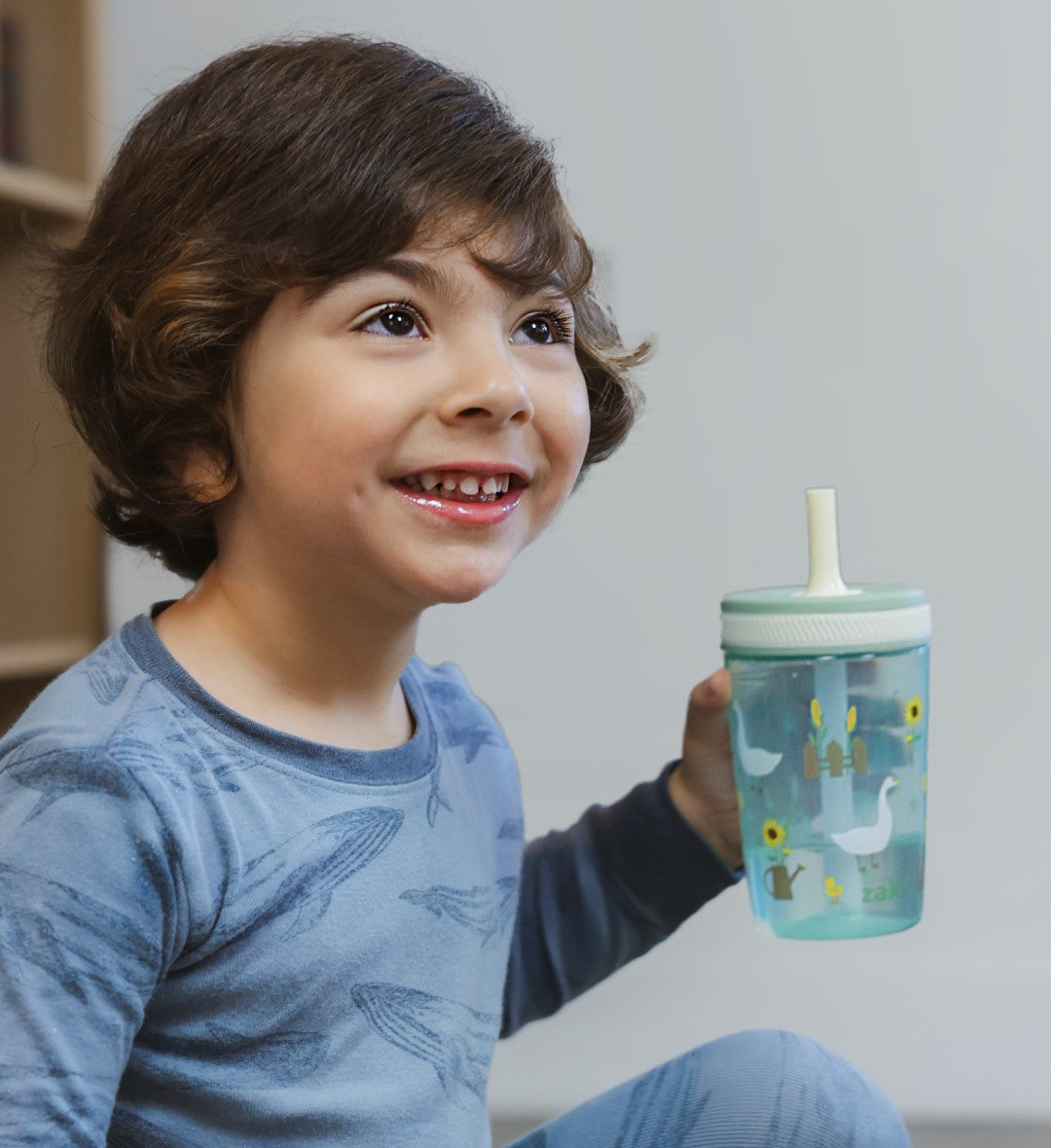 Child holding a blue Zak Designs sippy cup with a straw lid, smiling.