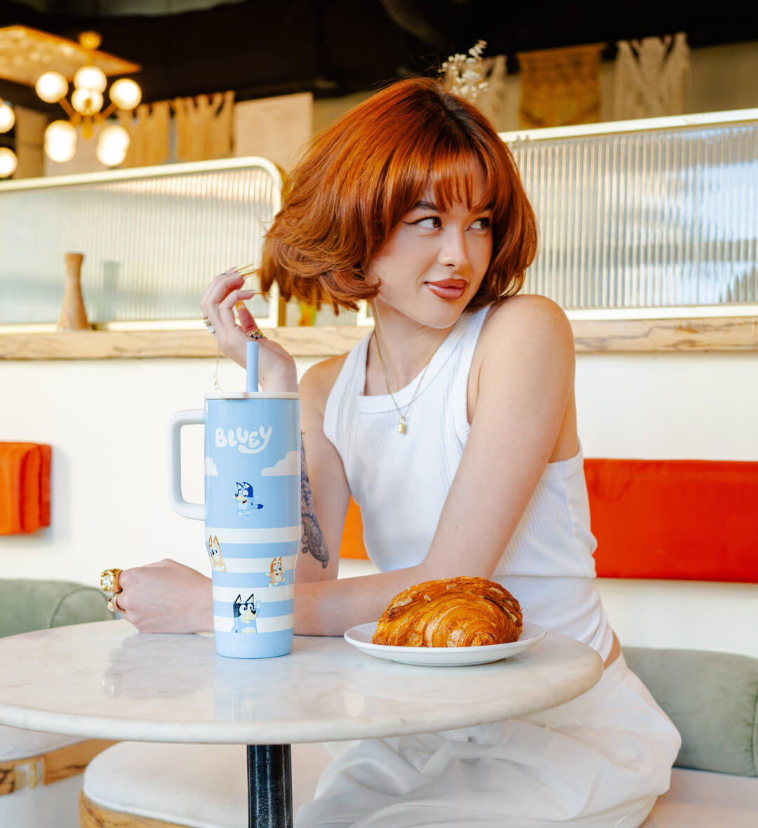 Woman sitting at a table with a Bluey tumbler and a pastry in a cafe.