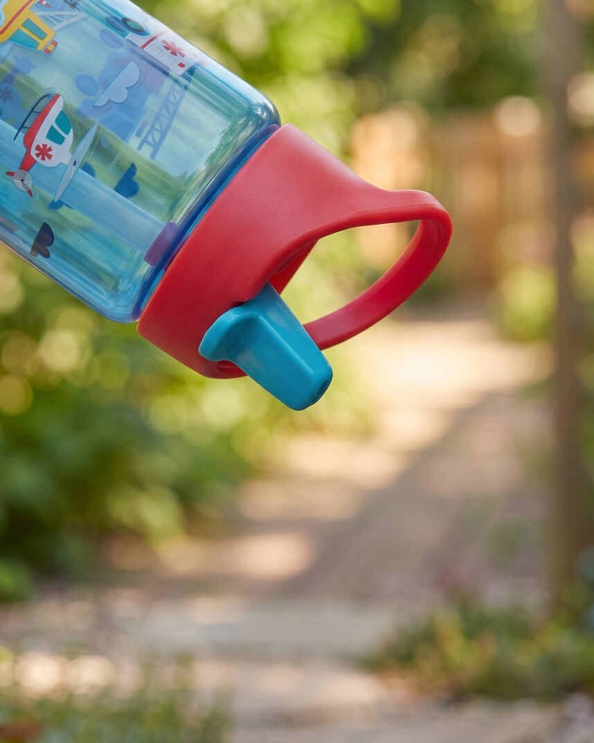 A close-up of a children's water bottle with a translucent blue body, red handle, and turquoise spout, photographed outdoors against a blurred garden background.