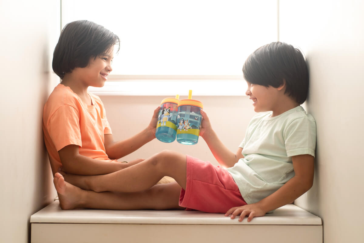 Two children sitting on a windowsill holding colorful zak Bluey cups.