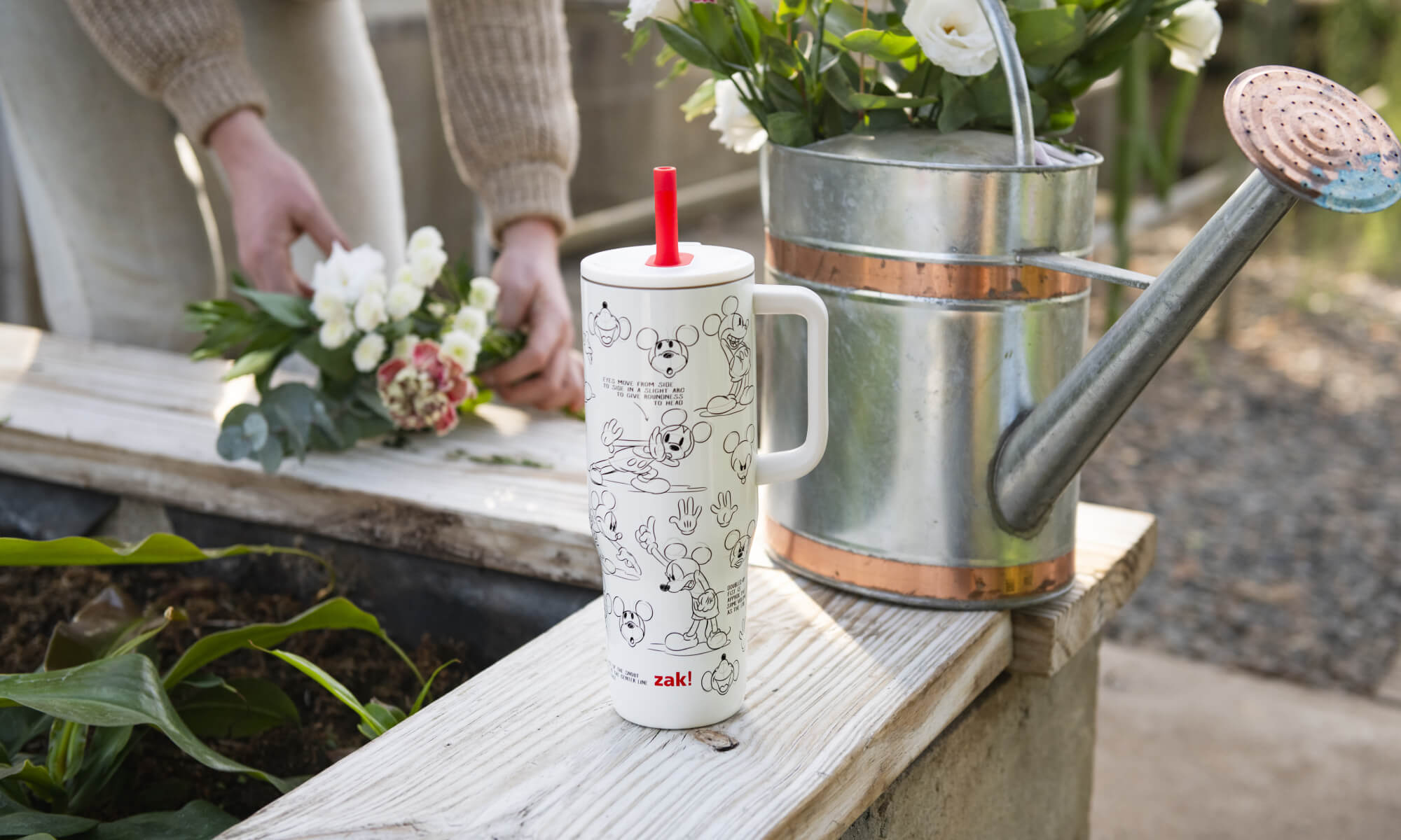 Zak Disney tumbler with a red straw lid on a wooden surface next to a watering can and flowers.