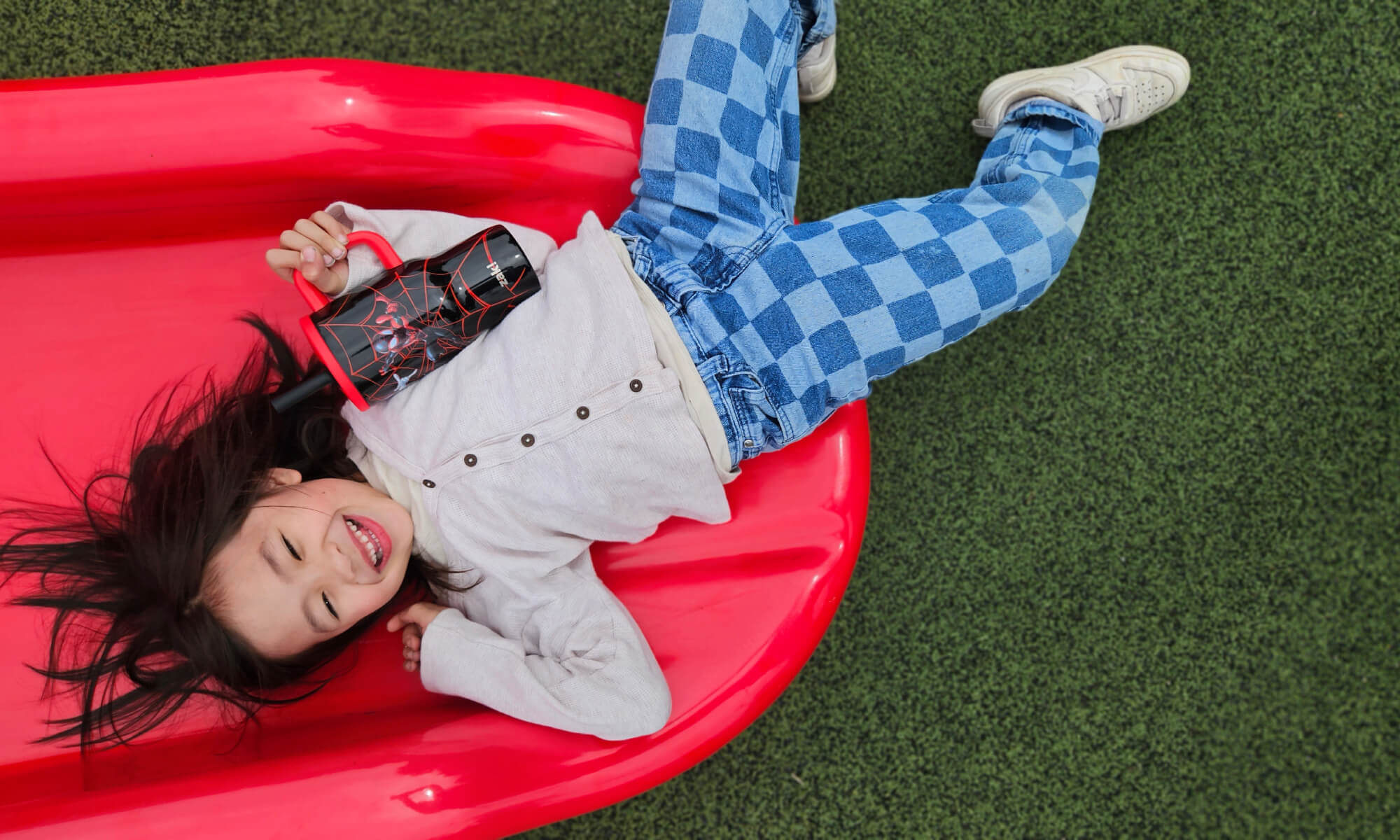 Child sliding down a red slide on a playground with Spider-Man tumbler