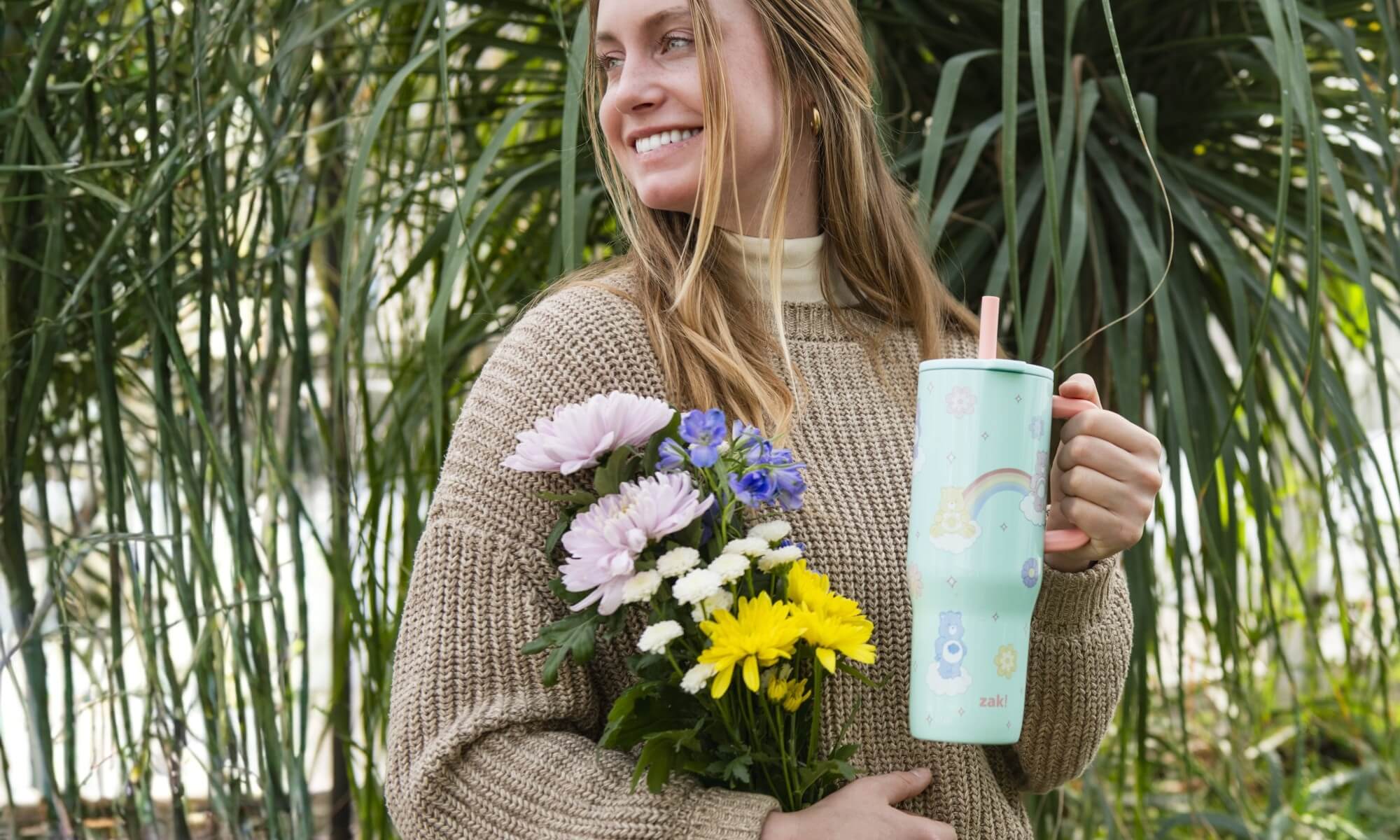 Woman holding a floral arrangement and a zak tumbler with a plant-themed design, standing in front of green foliage.