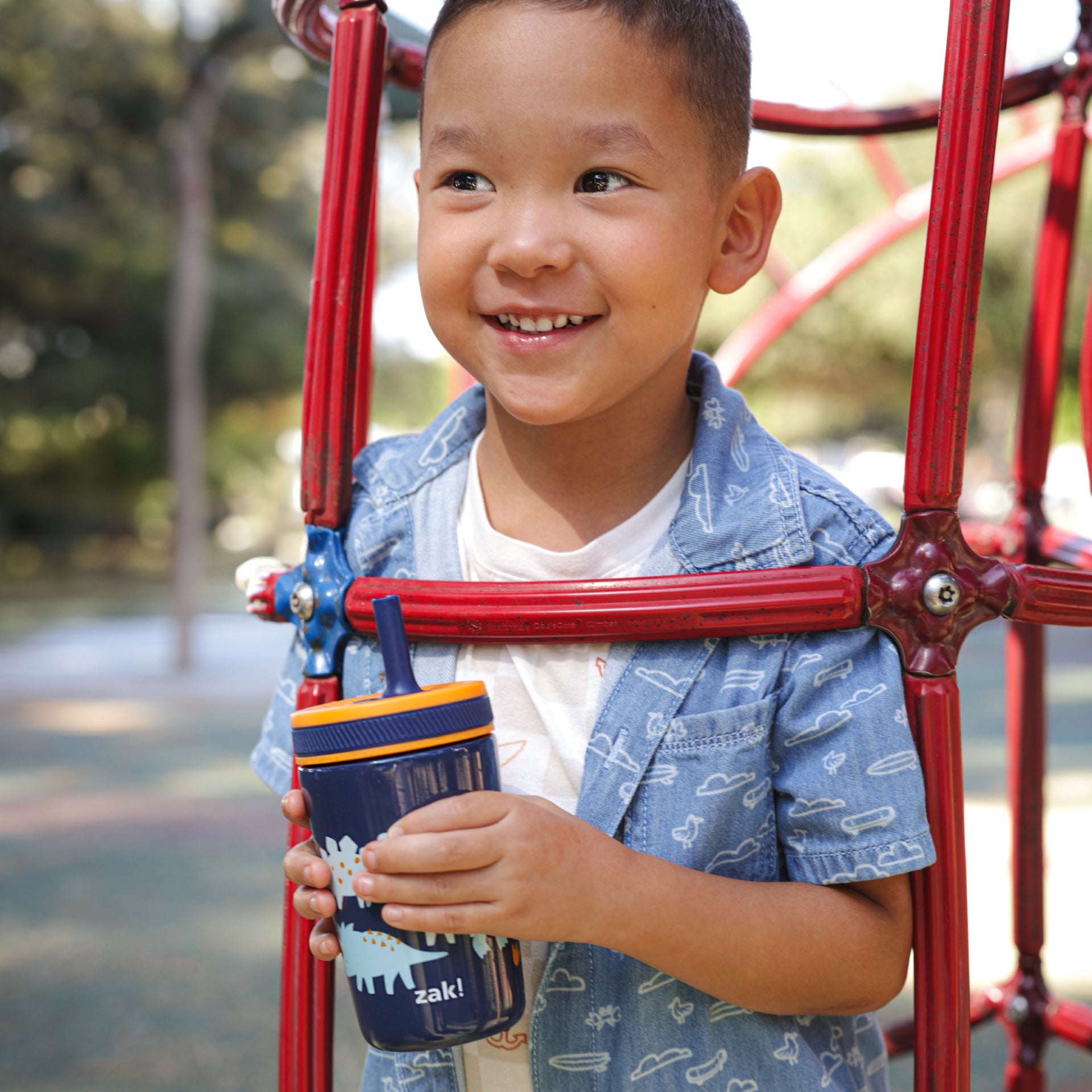 Child holding a blue tumbler with a red playground structure in the background