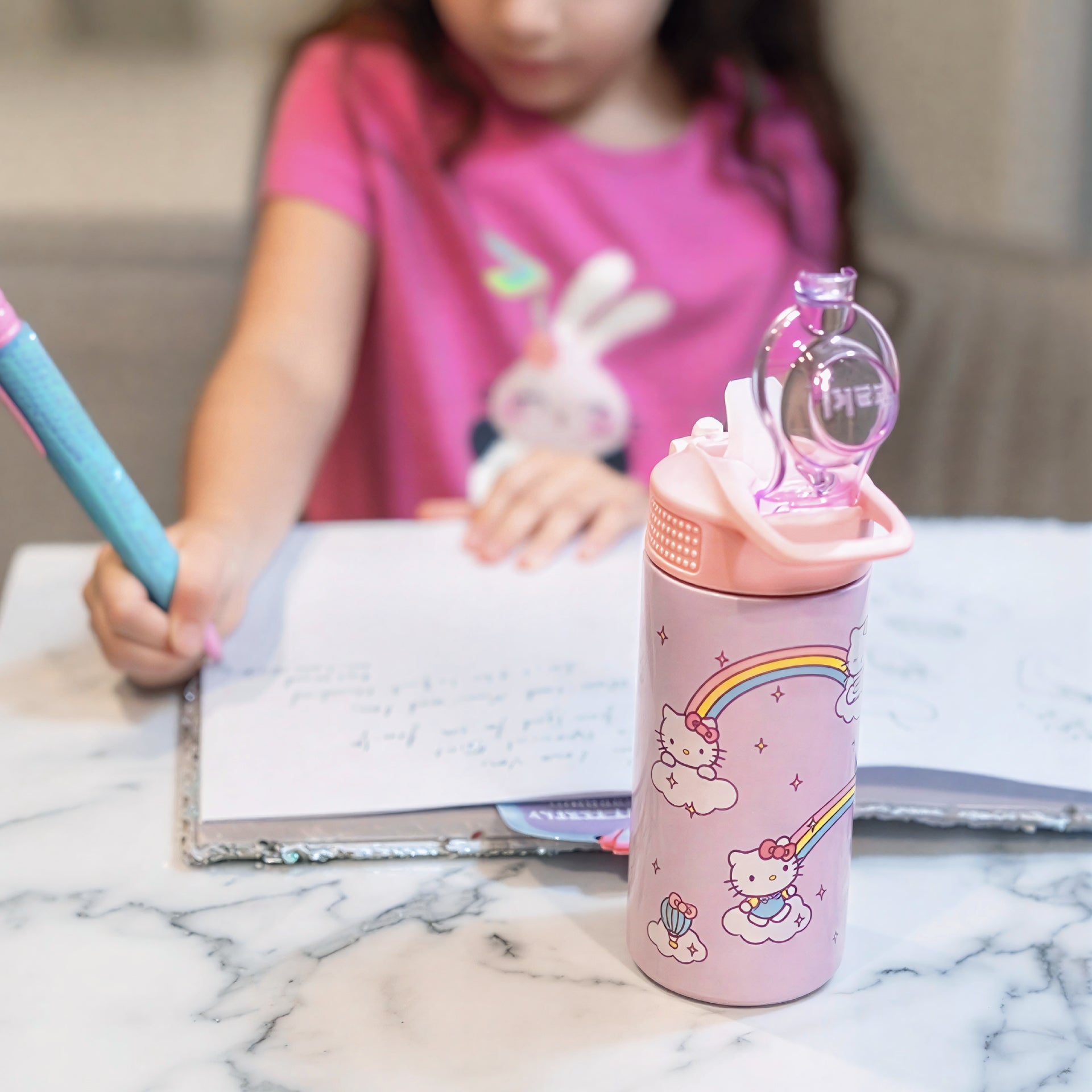 Pink Hello Kitty water bottle with character design on a marble surface, child writing in the background.