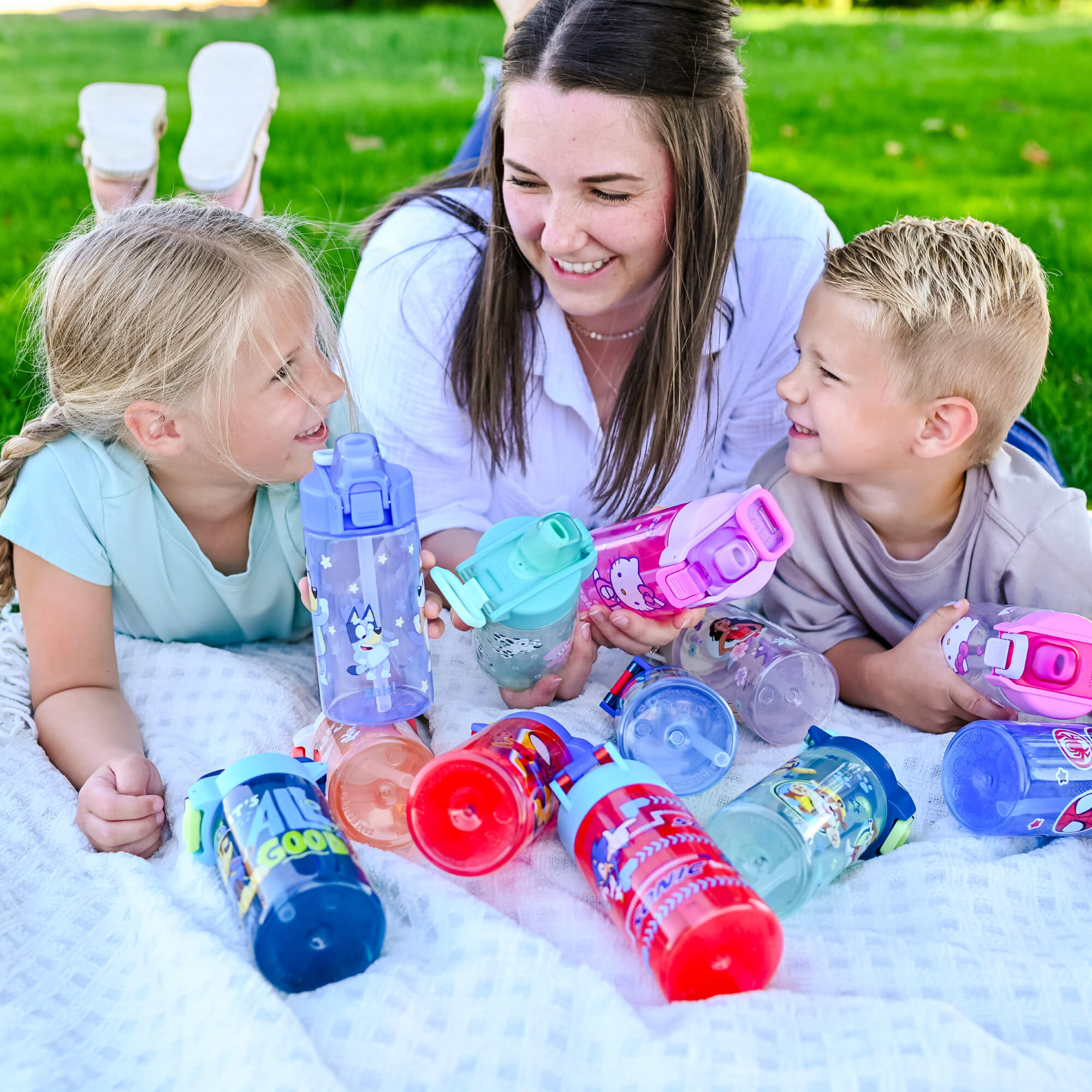 Happy mother and children laying in grass with blanket and colorful zak water bottles