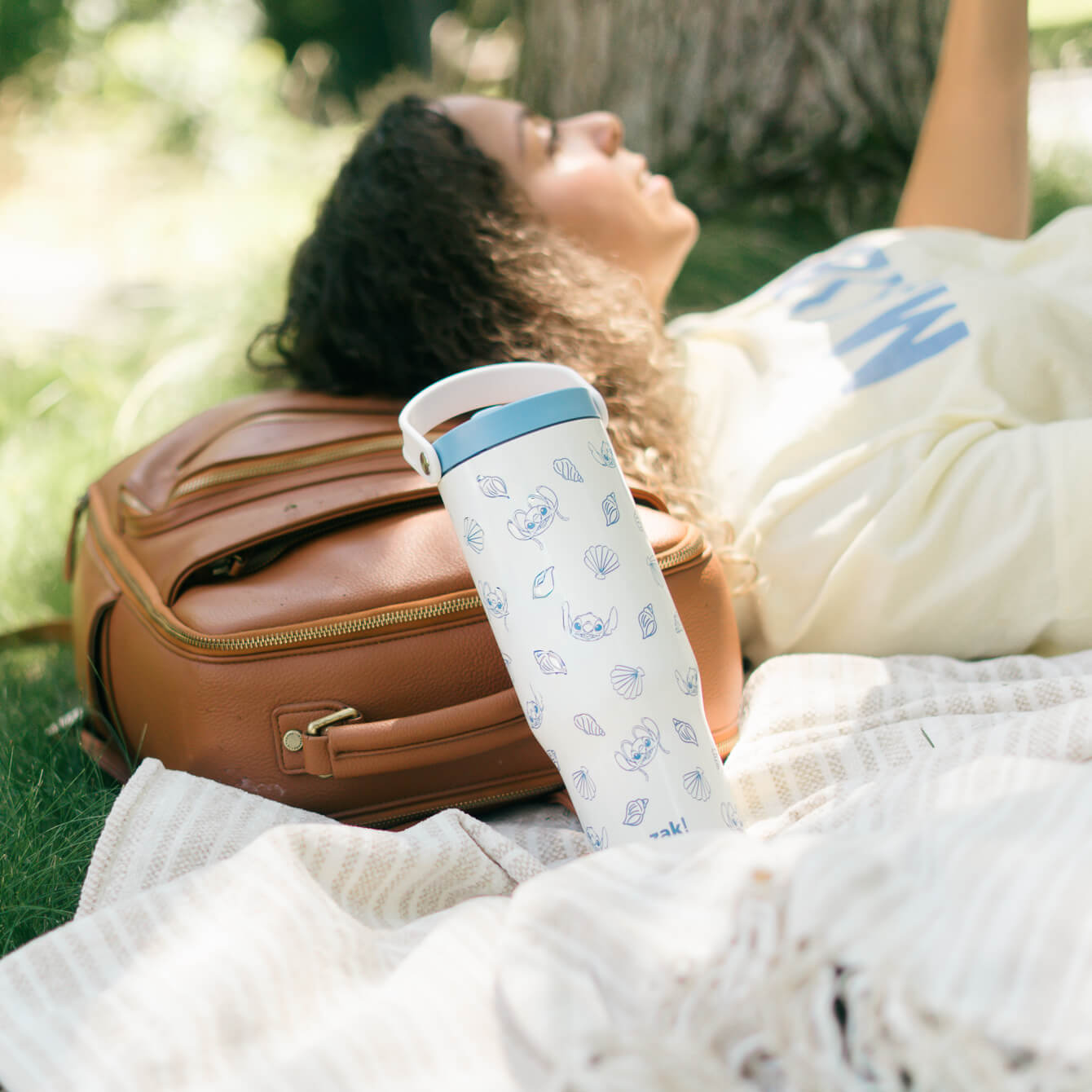 White tumbler leaning on a brown bag while woman in background is reading a book outdoor