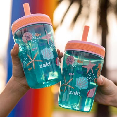 Two sea shells tumblers with pink lids and straw holders, featuring sea-themed designs, held by a person against a blurred outdoor background.