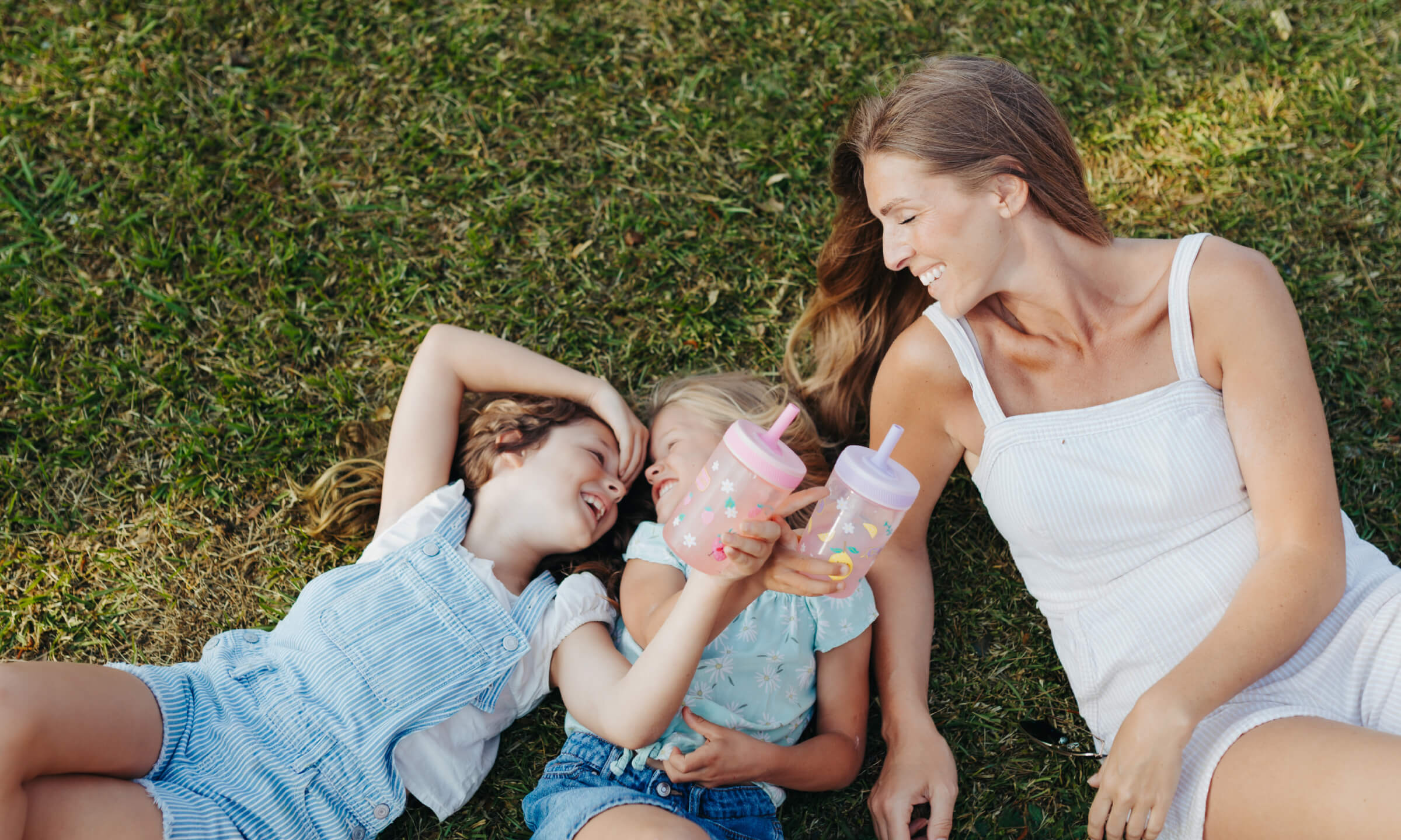 A woman and two young girls relax on a grassy lawn, sharing pink and blue sippy cups. The woman wears a white tank top and shorts, while the girls are in light blue and light green outfits.