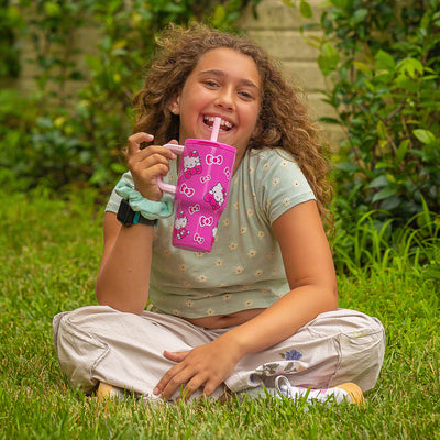 Child sitting on grass holding a pink zak Hello Kitty tumbler with a straw
