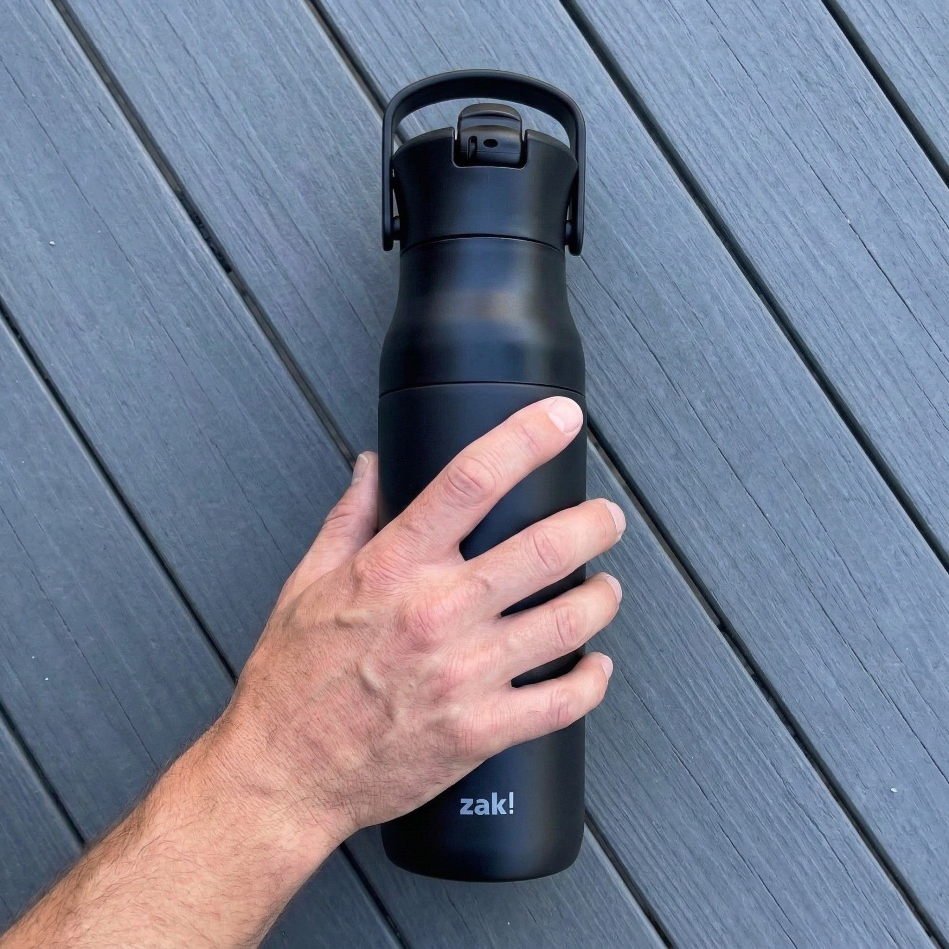 Hand holding a black water bottle with 'zak!' branding on a wooden surface