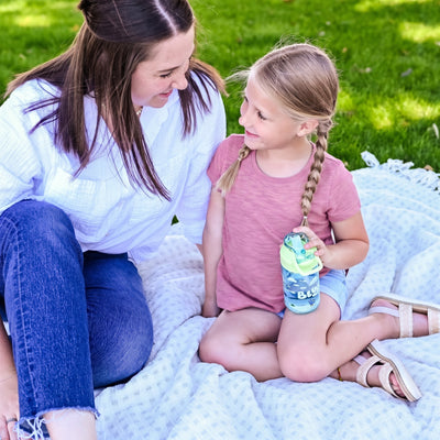A woman and a young girl sitting on a white blanket on grass, with the girl holding a blue and green water bottle. The woman is wearing a white shirt and blue jeans, and the girl is wearing a pink t-shirt and denim shorts.